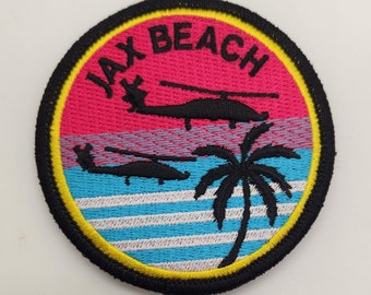 May include: A round, black and yellow embroidered patch with the text "JAX BEACH" in white. The patch features a palm tree and two helicopters in black against a pink, blue, and white striped background.