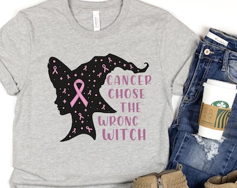 May include: A grey t-shirt with a black silhouette of a witch wearing a pink ribbon. The text "Cancer chose the wrong witch" is printed on the shirt.