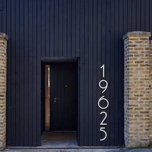 May include: A black wooden house with a brick wall and a black door. The house number 19625 is displayed on the wall.
