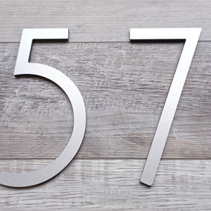 Pu&ograve; includere: Numeri civici in metallo argentato 57 su uno sfondo in legno.