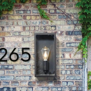 May include: Black metal house number plaque with the number 265 on a brick wall. A black metal light fixture with a clear glass bulb is mounted on the wall.
