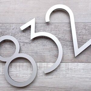 May include: Three silver metal house numbers, 8, 5, and 2, are displayed on a light wood background.
