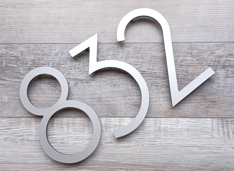 May include: Three silver metal house numbers, 8, 5, and 2, are displayed on a light wood background.