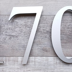 May include: Silver metal house numbers 170. The numbers are attached to a light gray wood surface.