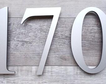 May include: Silver metal house numbers 170. The numbers are attached to a light gray wood surface.