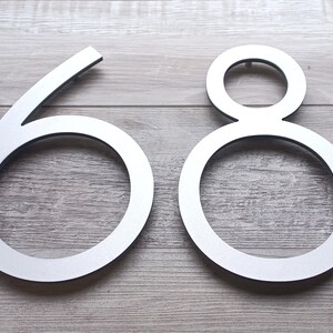 May include: Silver metal house numbers 68, with a modern, minimalist design. The numbers are attached to a light wood surface.