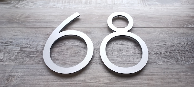 May include: Silver metal house numbers 68, with a modern, minimalist design. The numbers are attached to a light wood surface.