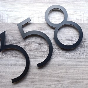 May include: Black metal house numbers 358 on a light wood background.