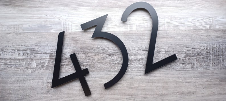 May include: Black metal house numbers 4, 3, and 2. The numbers are arranged in a random order on a light wood background.