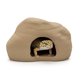 Op de afbeelding: Een beige reptielenverblijf in de vorm van een rots met een cirkelvormige opening. Een luipaardgekko met gele en zwarte vlekken zit erin. Het verblijf is ontworpen voor reptielen.