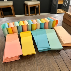 May include: A paper chain made of interlocking loops in pastel shades of peach, yellow, mint green, and light blue. Beside the chain are several neat stacks of rectangular paper strips in matching colors, arranged in rows on a wooden surface.