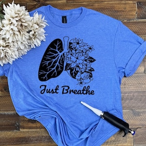 May include: A blue t-shirt with a black graphic of a lung with flowers growing out of it. The text "Just Breathe" is printed below the graphic.