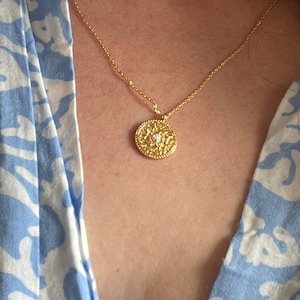 May include: A gold chain necklace with a round pendant featuring a delicate floral design and a small white stone in the center.