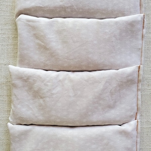 Puede incluir: Cuatro almohadas para ojos de lino blanco apiladas una encima de la otra. Las almohadas están cosidas con hilo marrón.