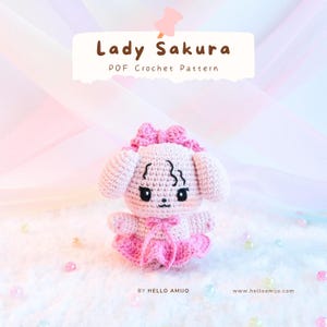 May include: A crocheted pink puppy doll with a bow on its head and a pink dress. The doll has black eyes and a small bow at the neck. The text "Lady Sakura PDF Crochet Pattern" is above the doll.