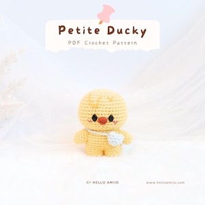 Può includere: Un'anatra gialla all'uncinetto con un piccolo accessorio a forma di cuore blu. L'anatra ha occhi neri e un becco arancione. Il testo sull'immagine recita "Petite Ducky PDF Crochet Pattern" e "By Hello Amijo".