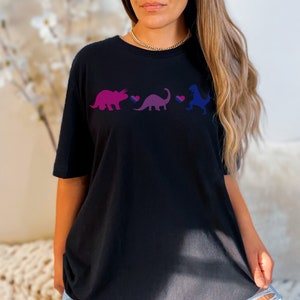 Pode incluir: Uma t-shirt preta de manga curta com uma estampa gráfica de três dinossauros em tons de rosa, roxo e azul. Entre cada dinossauro, há um pequeno ícone de coração com listras horizontais nas cores rosa, roxo e azul, dispostos em linha.