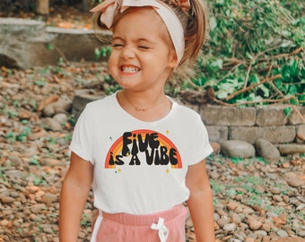 May include: A white short-sleeved t-shirt featuring a graphic of a multi-colored rainbow in shades of orange and yellow with the black text "Five is a vibe" centered in the middle, accompanied by small four-pointed star accents in yellow and blue.