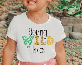 May include: A white short-sleeved t-shirt featuring the black text Young Wild and Three. The word Wild is printed in large block letters with a green and yellow animal print pattern. The shirt is paired with pink pants featuring a white drawstring.