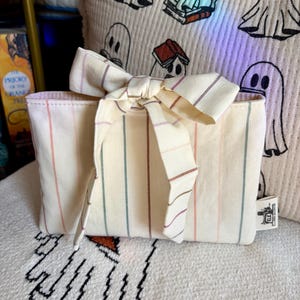 May include: A rectangular cream-colored fabric pouch featuring thin vertical stripes in muted shades of pink, green, and brown. The pouch is tied at the top with a matching fabric bow and includes a small white tag on the side with black text.