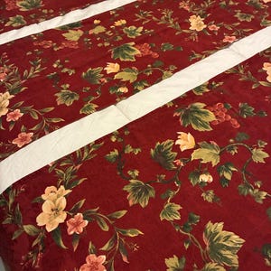 May include: A fabric runner featuring a deep red background with a repeating pattern of green leaves, light yellow flowers, and clusters of small red grapes. The runner is accented by two distinct, parallel white stripes running horizontally.