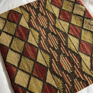 May include: Square decorative pillow with a geometric pattern. The design features diamond shapes in shades of gold, burgundy, and black. The pillow has a textured appearance, suggesting a woven fabric.