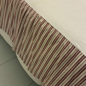 May include: A bed skirt with a cream-colored top and a striped lower section. The stripes are vertical, alternating between burgundy, cream, and light gray. The bed skirt is made of a soft fabric and has a gathered design.