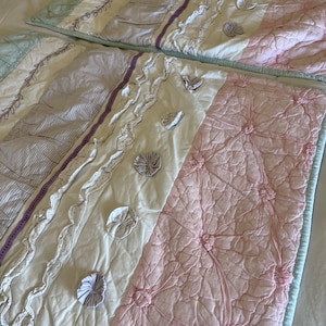 May include: Two decorative quilted bed pillows. The pillows feature a patchwork design with pastel colors, including light pink, white, and pale blue. The pillows have ruffled and floral accents, with a light blue border.