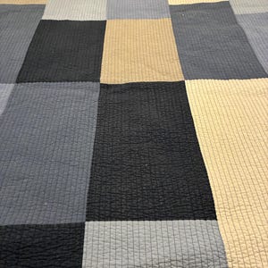 May include: A quilt featuring a grid pattern of rectangular fabric panels in shades of black, light blue, and beige. The surface of the quilt displays a consistent, fine linear quilted texture across all the colored sections of the fabric.