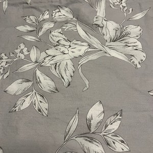 May include: A gray fabric featuring a repeating botanical print with white line-drawn flowers and leaves. The design showcases detailed illustrations of blossoms and foliage scattered across the surface, creating a monochromatic floral pattern.