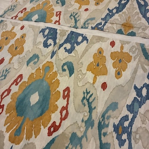May include: A fabric panel featuring an ikat pattern with large floral-inspired motifs in shades of blue, mustard yellow, and red on an off-white background. The textile displays a blurred, bleeding edge effect characteristic of the ikat technique.