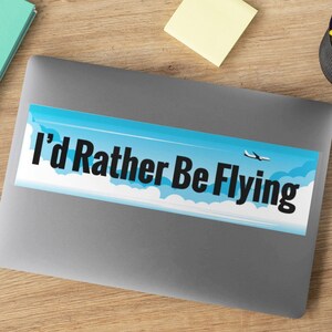 May include: A blue and white sticker with a white airplane flying in a blue sky with white clouds. The sticker says "I'd Rather Be Flying" in black text.