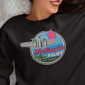 May include: Black sweatshirt featuring a colorful graphic of a multi-engine airplane inside a blue circle with the text "Multiengine Pilot" and "Fly Twins". The airplane has green and gray details, and a pink arrow points upwards.