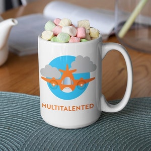 May include: White ceramic mug filled with colorful marshmallows. The mug features a cartoon airplane design in orange and blue, set against a blue circle and gray clouds. The word "MULTITALENTED" is printed below the design.