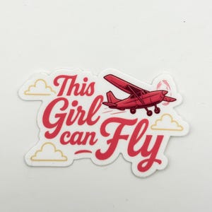 May include: A sticker with the text "This Girl can Fly" in red script, accompanied by a red airplane illustration. The design includes yellow clouds and a white background.