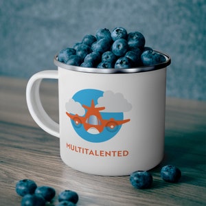May include: A white enamel mug with a silver rim, filled with blueberries. The mug features a cartoon airplane design in orange and blue, with the word "MULTITALENTED" in orange. The mug sits on a wooden surface.