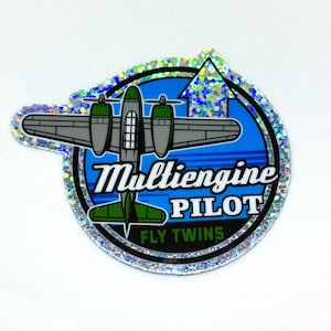 May include: A silver holographic sticker with a blue background and a cartoon airplane. The text "Multiengine Pilot Fly Twins" is printed on the sticker.