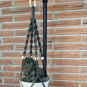 May include: A green macrame plant hanger with wooden beads hangs from a black metal stand. A white ceramic pot with a cactus plant is inside the hanger.