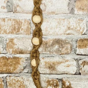 Puede incluir: Un llavero de macramé marrón con tres cuentas de madera. El llavero está colgado de un anillo plateado sobre un fondo de pared de ladrillo.