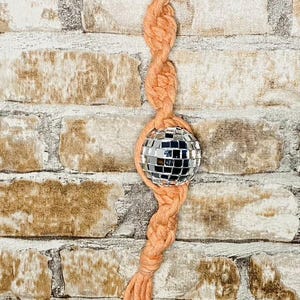 May include: A peach-colored macrame keychain with a silver disco ball charm. The keychain is hanging from a silver ring.