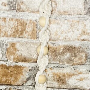 May include: A white macramé plant hanger with three wooden beads. The hanger has a silver metal ring at the top for hanging. The macramé is made of thick, twisted cotton rope.