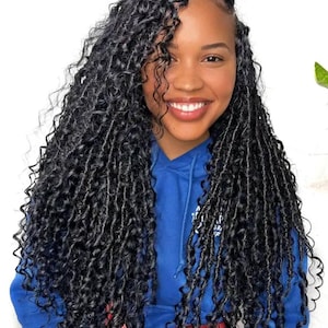 May include: A smiling person with long, dark, curly hair styled in a locs hairstyle. The person is wearing a blue hoodie and a red fanny pack. The background is a plain white wall.