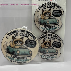 May include: Car coasters with a Grumpy Cat design. The coasters feature the cat in a light blue car, giving the middle finger, with text that reads "Thanks for driving so close." and "I appreciate your confidence in my reflexes."