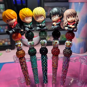 May include: A set of five decorative pens featuring character figurines. Each pen has a different color of sparkling rhinestones on the barrel. The figurines are of characters from a popular fantasy series. The pens are displayed in a clear plastic stand.