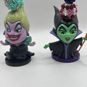 May include: Two cartoon figurines of Disney villains, Ursula and Maleficent. Ursula is purple with green hair and a black octopus base. Maleficent is green with black horns and a purple base. Both have decorative beaded toppers.