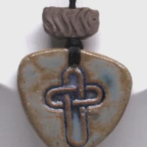 May include: A ceramic pendant featuring a triangular shape with a glazed blue and tan finish. A dark blue embossed cross design is centered on the surface. The pendant hangs from a black cord with a textured, rectangular brown bead above it.