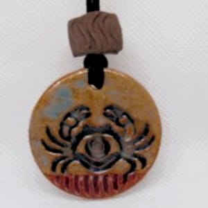 May include: A ceramic pendant necklace with a brown cord and a round, brown and gold pendant featuring a black crab design.