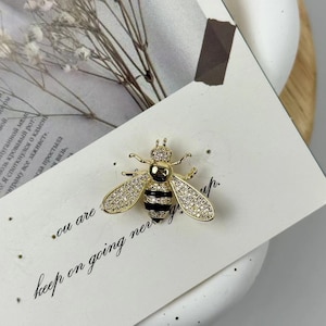 May include: A gold bee brooch with black stripes and clear rhinestones. The bee is on a white background with the text "you are keep on going never give up."
