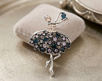 May include: Silver-toned ballerina brooch with a tutu adorned with teal and lavender crystals. The dancer holds a blue crystal. The brooch is displayed on a gray velvet cushion, suggesting a jewelry or accessory theme.