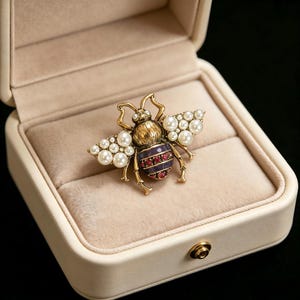 Peut inclure: Broche dorée en forme d'abeille, ornée de perles sur les ailes et de pierres rouges sur l'abdomen. Elle est présentée dans un écrin crème avec un intérieur en velours, mettant en valeur le design et le savoir-faire.
